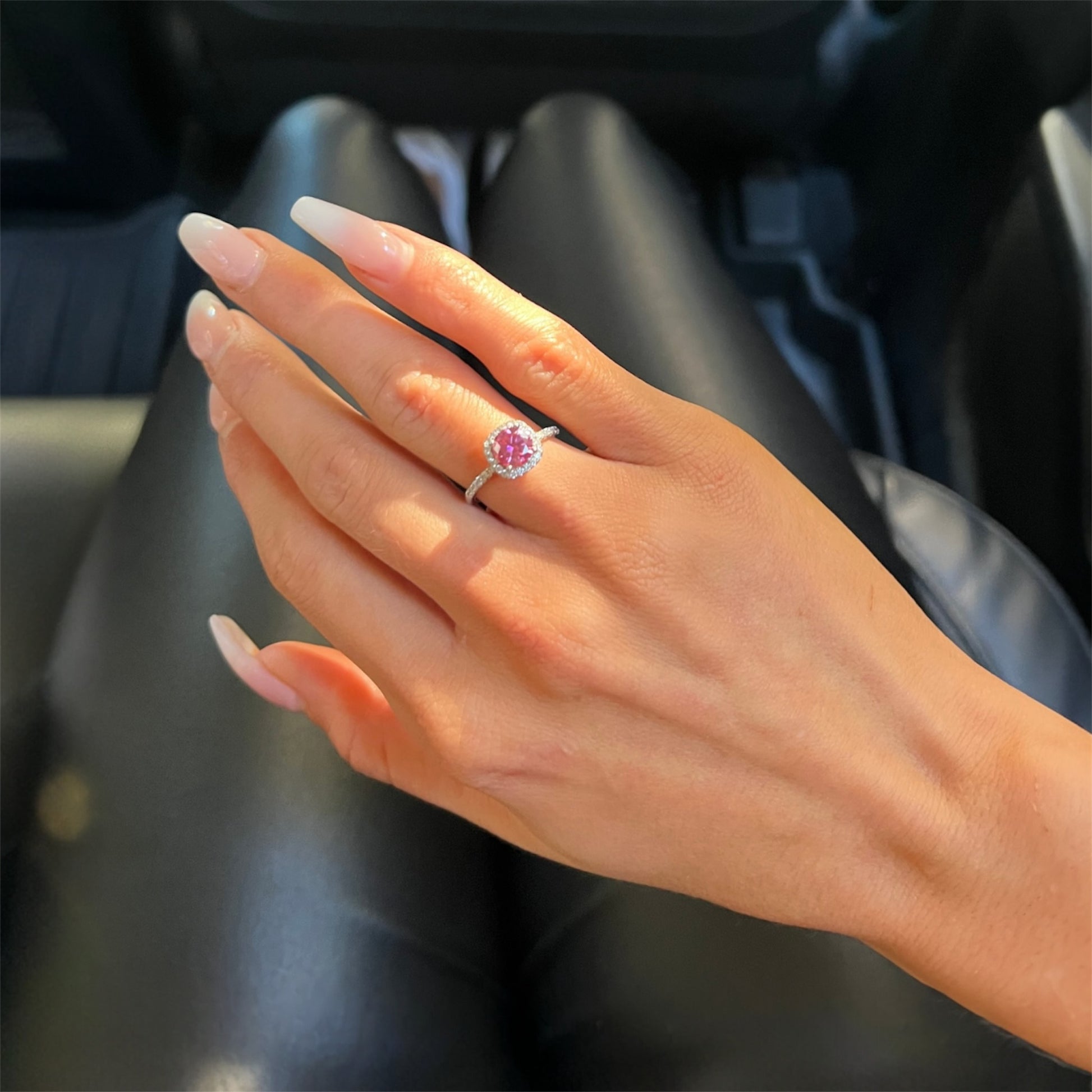 Hand wearing a 1 carat pink Moissanite halo ring on a black leather surface